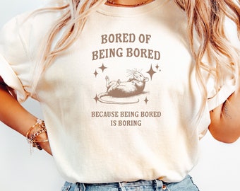 May include: Cream-colored t-shirt with the text "BORED OF BEING BORED" and a graphic of a relaxed rat. The phrase "BECAUSE BEING BORED IS BORING" is printed below the graphic. The model is wearing light blue jeans.