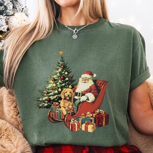 May include: A moss green t-shirt featuring a Christmas graphic. The graphic depicts Santa Claus in a sleigh with a golden retriever and a decorated Christmas tree. Presents are at the base of the sleigh. The shirt has rolled-up sleeves.