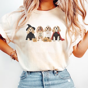 May include: A white t-shirt with a graphic of four dogs dressed in Halloween costumes. The dogs are wearing a witch hat, a trick or treat bucket, a mummy costume, and a vampire cape. The dogs are sitting in front of pumpkins.