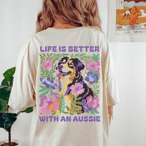 May include: Ivory t-shirt with a graphic of an Australian Shepherd dog surrounded by flowers. The text "Life is better with an Aussie" is printed on the back of the shirt.