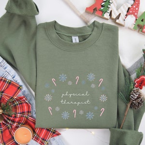 May include: A green sweatshirt with a white snowflake and candy cane design and the text "physical therapist" in white.