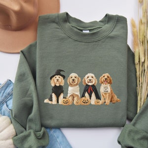 May include: Green sweatshirt featuring a Halloween-themed graphic of four dogs dressed in costumes. The dogs are wearing a witch hat, mummy bandages, a cape, and a "Trick or Treat" bib, each sitting next to a carved pumpkin.