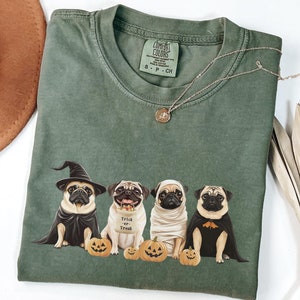 May include: Green t-shirt with a graphic of four pugs dressed up in Halloween costumes. The pugs are wearing a witch hat, a "trick or treat" sign, a mummy costume, and a bat costume. The pugs are sitting in front of pumpkins.