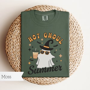 May include: A green t-shirt with a ghost wearing sunglasses and a witch hat holding a cup of coffee. The text on the shirt reads "Hot Ghoul Summer".