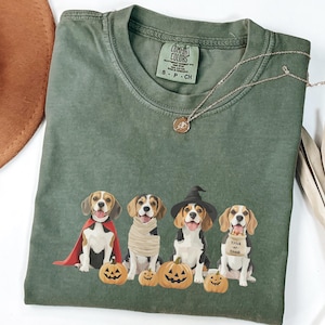 May include: Olive green t-shirt featuring a graphic of four beagles dressed in Halloween costumes, including a cape, mummy wrap, witch hat, and a "Trick or Treat" bib. The shirt also has jack-o'-lanterns. A gold necklace is draped across the shirt.