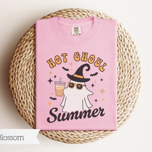 May include: Pink t-shirt with a graphic of a ghost wearing a witch hat and sunglasses, holding a cup of coffee. The text "Hot Ghoul Summer" is printed on the shirt.