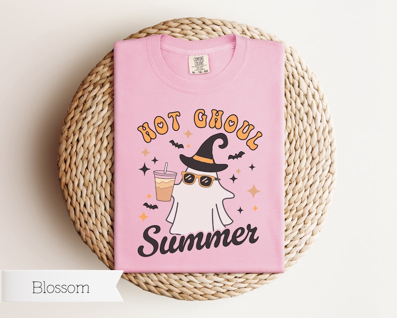 May include: Pink t-shirt with a graphic of a ghost wearing a witch hat and sunglasses, holding a cup of coffee. The text "Hot Ghoul Summer" is printed on the shirt.