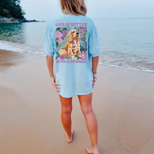 Op de afbeelding: Een lichtblauw t-shirt met een bloemenpatroon en een gouden spaniel-hond. De tekst op het shirt luidt "Life is better with a spaniel."