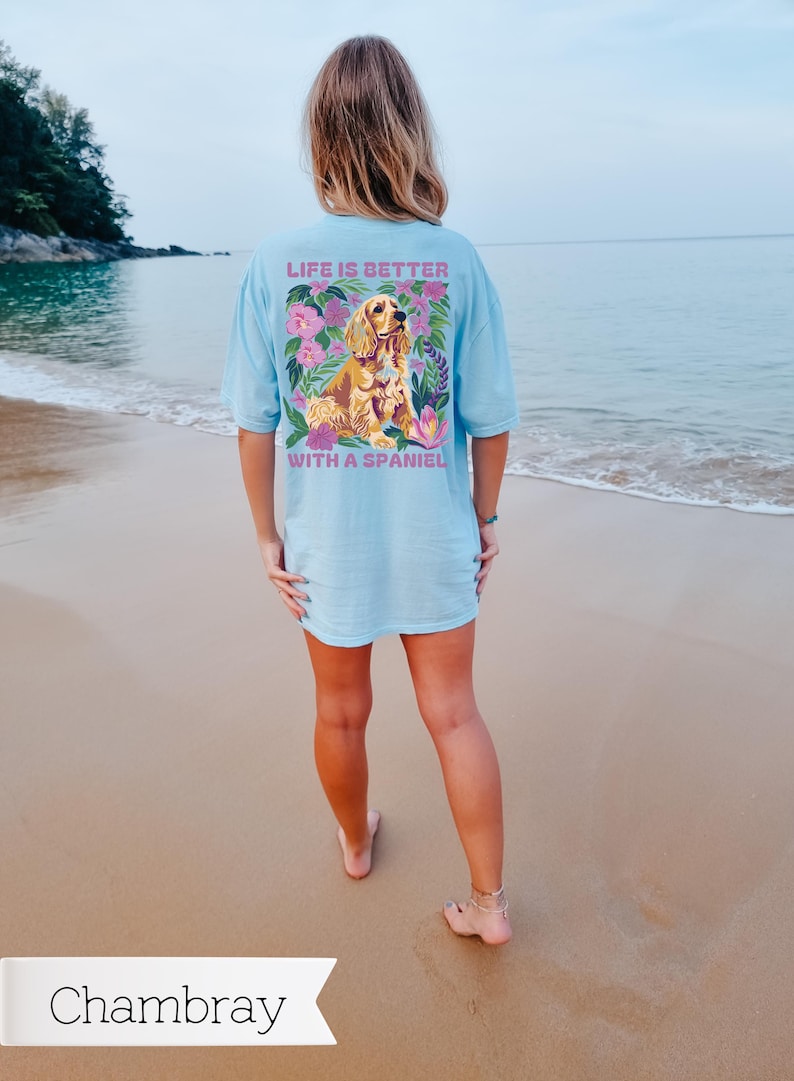 Op de afbeelding: Een lichtblauw t-shirt met een bloemenpatroon en een gouden spaniel-hond. De tekst op het shirt luidt "Life is better with a spaniel."