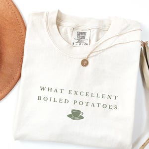 May include: A beige t-shirt with the text "What excellent boiled potatoes" and a small green illustration of a teacup and saucer.