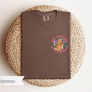 Op de afbeelding: Espresso bruin t-shirt met een afbeelding van een gouden spaniel in een bloemenkrans. De tekst "Life is better with a spaniel" is in roze rond de krans gedrukt.