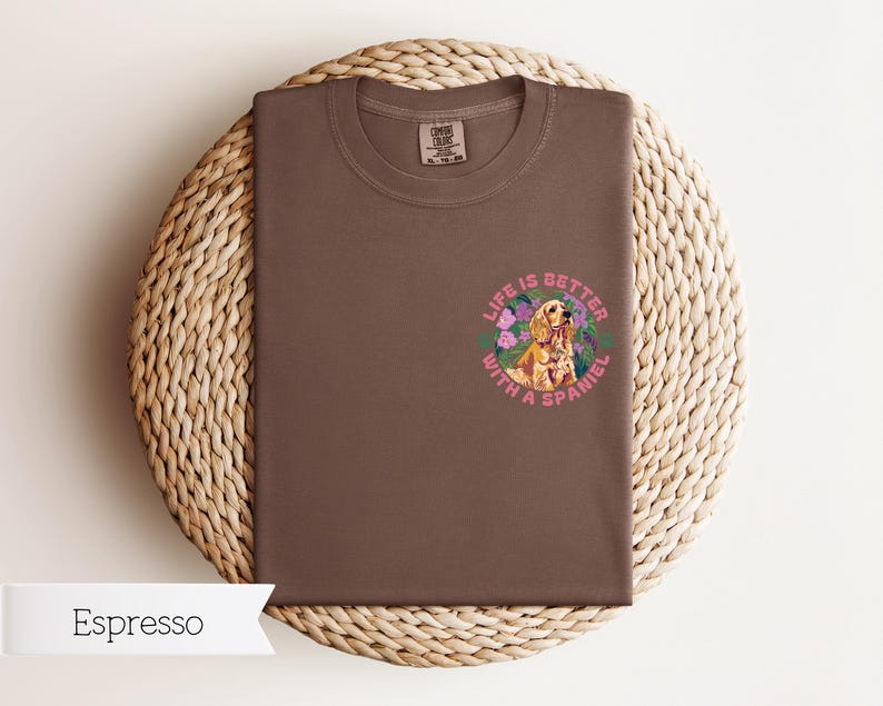 Op de afbeelding: Espresso bruin t-shirt met een afbeelding van een gouden spaniel in een bloemenkrans. De tekst "Life is better with a spaniel" is in roze rond de krans gedrukt.