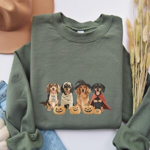 May include: Green sweatshirt featuring a Halloween-themed design. The graphic depicts four dachshund dogs dressed in costumes, including a mummy, witch, and vampire, with carved pumpkins. The phrase "Trick or Treat" is on one dog's costume.