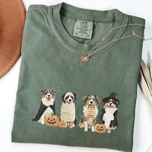 May include: Green t-shirt with a graphic of three Australian Shepherd dogs dressed up in Halloween costumes. The dogs are wearing a cape, a mummy costume, and a witch hat. The text "Trick or Treat" is on a bag held by one of the dogs.