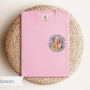 Op de afbeelding: Roze T-shirt met een afbeelding van een spaniel-hond met bloemen en de tekst "Life is better with a spaniel".