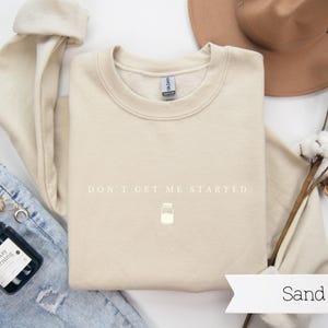 May include: A beige sweatshirt with the text "DON'T GET ME STARTED" in white lettering. A small white jar icon is below the text. The sweatshirt is laid flat on a white surface with a pair of blue jeans, a candle, and a hat.