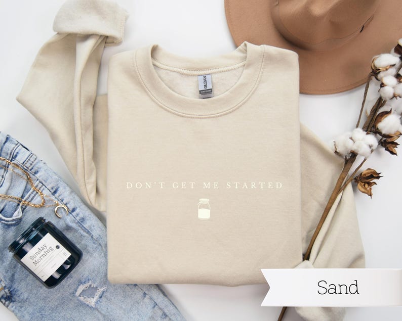 May include: A beige sweatshirt with the text "DON'T GET ME STARTED" in white lettering. A small white jar icon is below the text. The sweatshirt is laid flat on a white surface with a pair of blue jeans, a candle, and a hat.