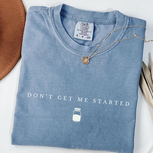 May include: A light blue t-shirt with the text "DON'T GET ME STARTED" in white lettering. A white outline of a milk jar is below the text.