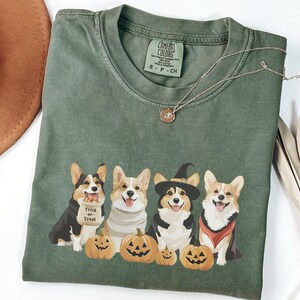 May include: Green t-shirt with a graphic of four corgis dressed in Halloween costumes. The corgis are holding pumpkins and the text "Trick or Treat" is on the shirt.