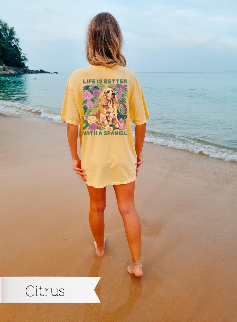 Op de afbeelding: Een geel T-shirt met een afbeelding van een hond in een bloemenontwerp. De tekst op het shirt luidt "Life is better with a spaniel."
