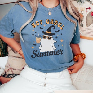 May include: A blue jean t-shirt with a white ghost wearing sunglasses and a witch's hat holding a cup of iced coffee. The text on the shirt reads "Hot Ghoul Summer".