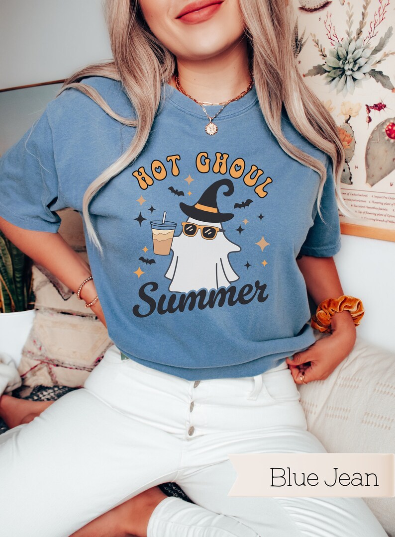 May include: A blue jean t-shirt with a white ghost wearing sunglasses and a witch's hat holding a cup of iced coffee. The text on the shirt reads "Hot Ghoul Summer".
