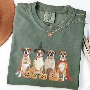 May include: Green t-shirt with a graphic of four boxer dogs dressed in Halloween costumes. The dogs are wearing a witch hat, a mummy costume, a vampire costume, and a superhero costume. The text "Trick or Treat" is printed on the shirt.