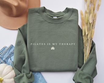 Puede incluir: Una sudadera verde oliva con el texto blanco "PILATES IS MY THERAPY" y un gráfico de flor de loto. La imagen incluye un sombrero marrón, vaqueros azules y plantas decorativas secas.