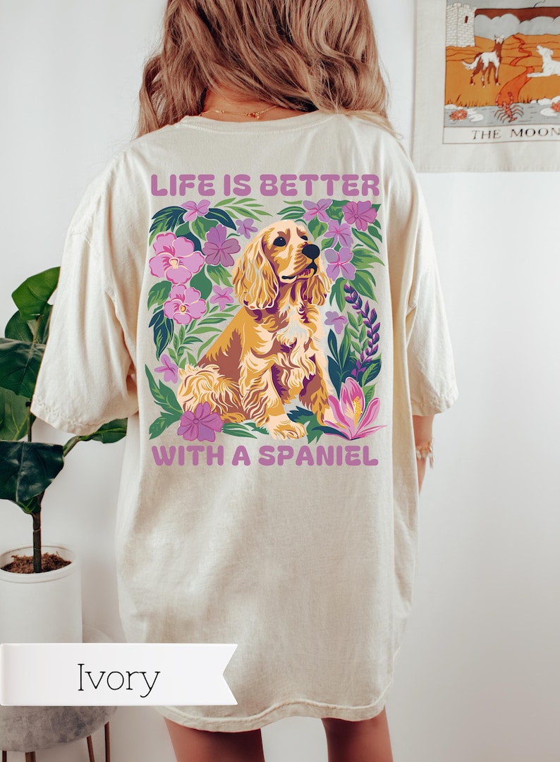 Op de afbeelding: Een ivoorwit T-shirt met een afbeelding van een gouden cocker spaniel die in een bloemstuk zit. De tekst "Life is better with a spaniel" staat op de achterkant van het shirt.