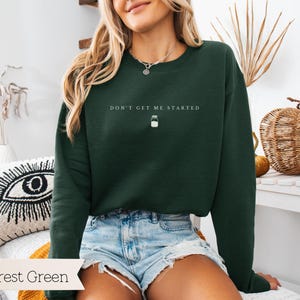 May include: Forest green sweatshirt with the text "Don't get me started" and a small jar icon. The sweatshirt is being worn by a person with long brown hair.