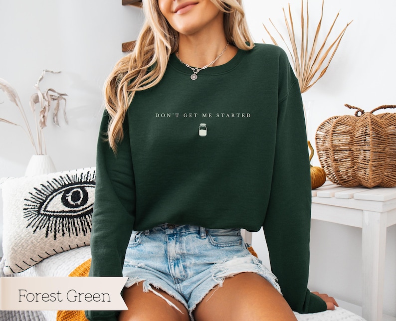 May include: Forest green sweatshirt with the text "Don't get me started" and a small jar icon. The sweatshirt is being worn by a person with long brown hair.