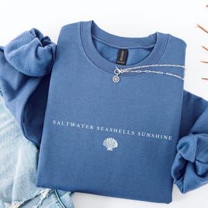 May include: A blue sweatshirt with the text "Saltwater Seashells Sunshine" and a white seashell graphic. The sweatshirt is folded and laid on a white surface with a pair of blue jeans.