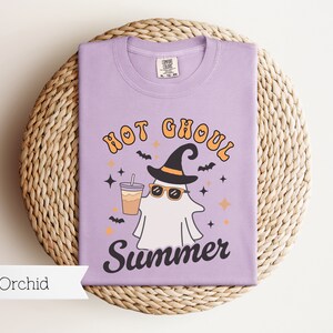 May include: A purple t-shirt with a ghost wearing a witch hat and sunglasses holding a cup of coffee. The text on the shirt reads "Hot Ghoul Summer".