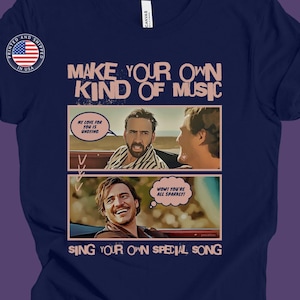 Può includere: Una t-shirt blu navy con una grafica in stile fumetto con Nicolas Cage. La grafica dice "Make your own kind of music" e "Sing your own special song".