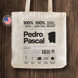 Puede incluir: Una bolsa de tela blanca con un diseño en blanco y negro que presenta el texto "100% Papá 100% Niña" y "Pedro Pascal". El diseño también incluye una silueta de una cabeza de persona y un código de barras.