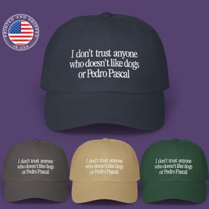 May include: A navy blue baseball cap with the text "I don't trust anyone who doesn't like dogs or Pedro Pascal" in white. Additional caps in gray, tan, and green are also shown. A circular graphic with the American flag and the text "Printed and Shipped in USA" is in the upper left.