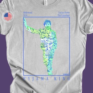 May include: A light gray t-shirt with a blue border around a graphic of a person in a green and blue patterned shirt and pants. The text "Hollywood 2024" is at the top of the graphic and "Lucian Flores The Uninvited" is at the top right of the graphic. The text "PYJAMA KING" is at the bottom of the graphic.