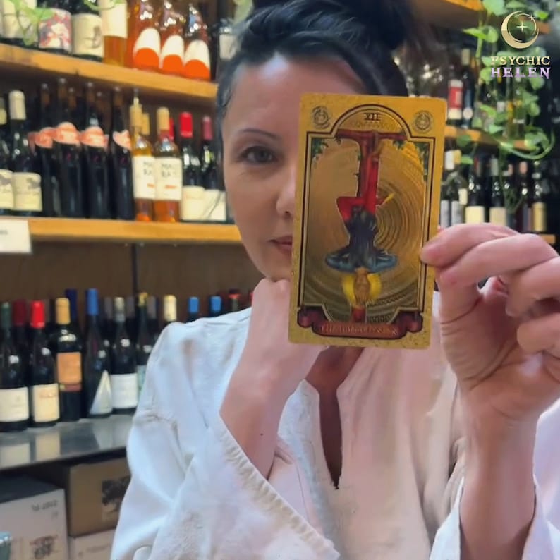 Pu&ograve; includere: Una carta dei tarocchi dorata raffigurante una figura capovolta con abiti rossi e blu, circondata da linee vorticose. La carta presenta il numero romano XII e del testo. Lo sfondo include scaffali di bottiglie di vino.