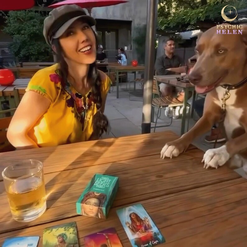 May include: A woman wearing a yellow shirt and a cap sits at a wooden table with a dog.  She has a glass of liquid and a deck of cards called "Light Seers" in front of her.  There are other cards on the table, including one that says "Queen of Cups".