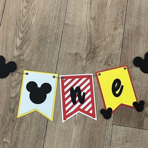 May include: A colourful banner with Mickey Mouse-themed decorations. The banner includes yellow, red, and black Mickey Mouse head shapes strung on twine. The pennants spell out "one" with a Mickey Mouse head on the first pennant.