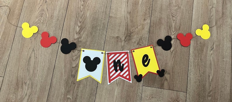 May include: A colourful banner with Mickey Mouse-themed decorations. The banner includes yellow, red, and black Mickey Mouse head shapes strung on twine. The pennants spell out "one" with a Mickey Mouse head on the first pennant.