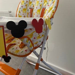 May include: A high chair with a white tray and orange details. The seat features a giraffe pattern. Mickey Mouse-shaped cutouts in black, red, and yellow are attached. A banner with the letter "e" is also attached to the chair.