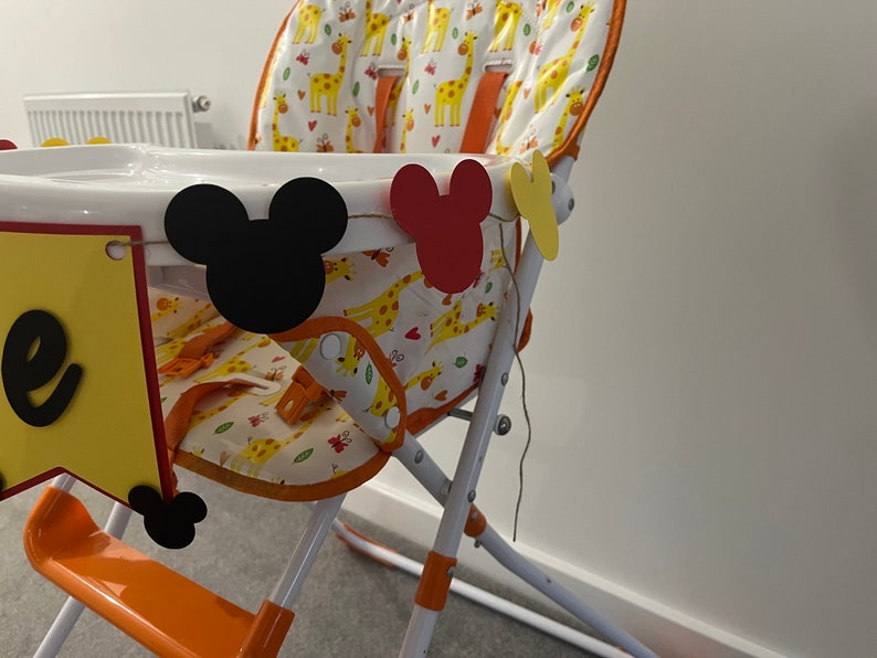 May include: A high chair with a white tray and orange details. The seat features a giraffe pattern. Mickey Mouse-shaped cutouts in black, red, and yellow are attached. A banner with the letter "e" is also attached to the chair.