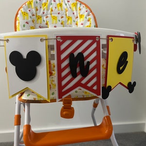 May include: A high chair decorated with a banner that says "one". The banner has three pennants: a white pennant with a black Mickey Mouse head, a red and white striped pennant with the letter "n", and a yellow pennant with the letter "e".
