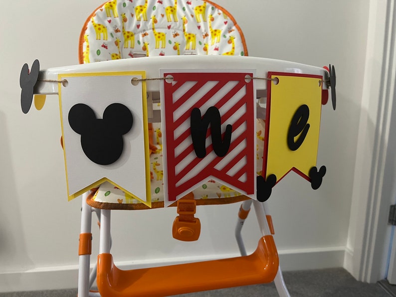 May include: A high chair decorated with a banner that says "one". The banner has three pennants: a white pennant with a black Mickey Mouse head, a red and white striped pennant with the letter "n", and a yellow pennant with the letter "e".