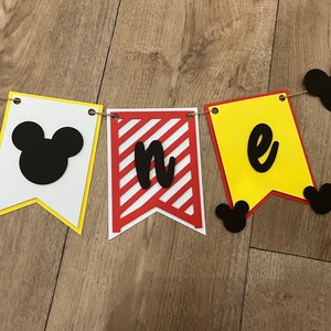 May include: A decorative banner spelling "one" with pennant flags in white, red, and yellow. The flags feature black Mickey Mouse silhouettes and lettering, strung together with twine for a birthday celebration.
