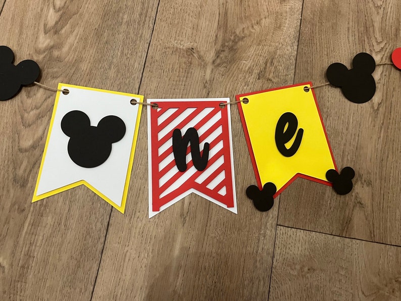 May include: A decorative banner spelling "one" with pennant flags in white, red, and yellow. The flags feature black Mickey Mouse silhouettes and lettering, strung together with twine for a birthday celebration.