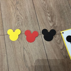 May include: A decorative banner with Mickey Mouse head shapes in yellow, red, and black, strung on twine. A white pennant with a black Mickey head is also included, ideal for a themed party or celebration.