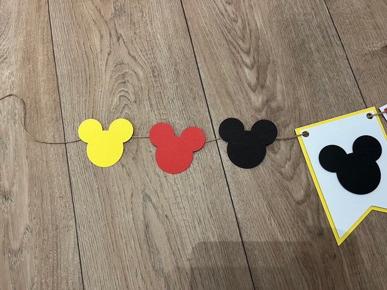 May include: A decorative banner with Mickey Mouse head shapes in yellow, red, and black, strung on twine. A white pennant with a black Mickey head is also included, ideal for a themed party or celebration.