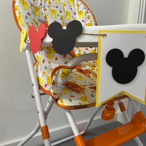 May include: A high chair with a giraffe-patterned seat and orange accents. A banner with Mickey Mouse head cutouts and pennants hangs across the front. The chair is white with orange and yellow details.